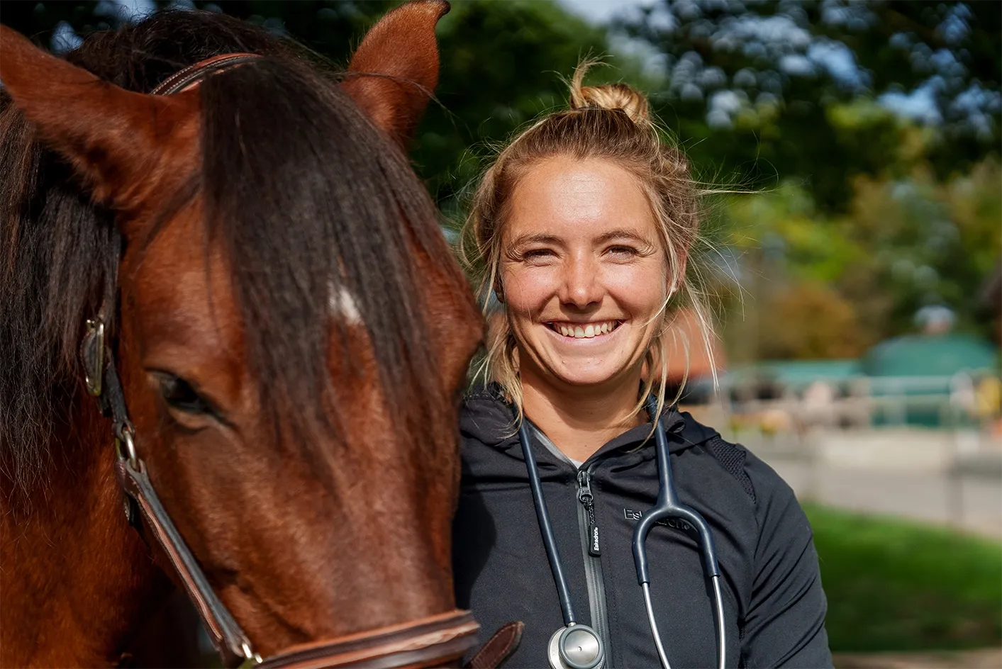 Tierärztin Jaqueline Göhle mit Stethoskop steht neben braunem Pferd im Freien