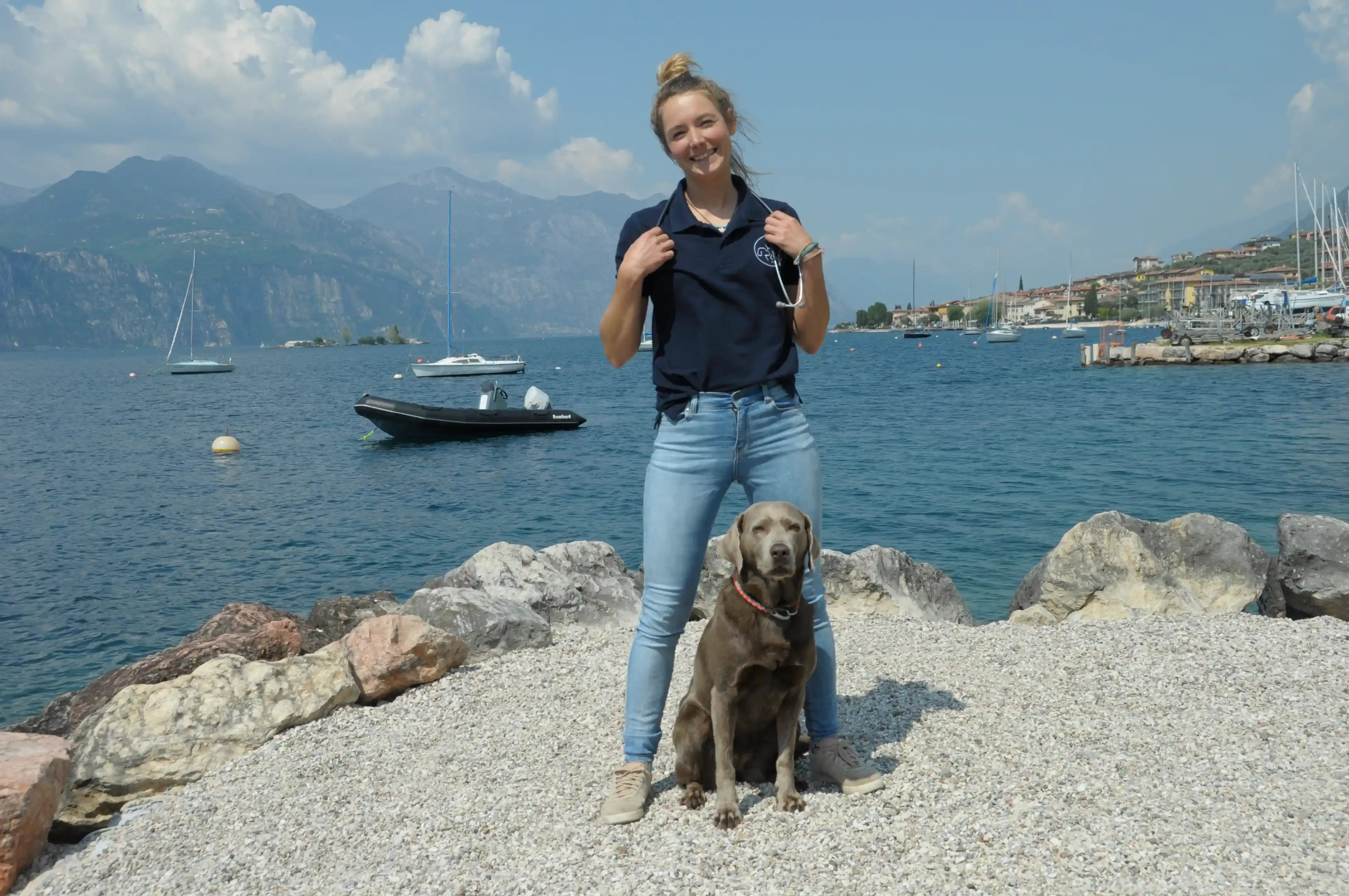 Person steht auf Kiesstrand am Gardasee mit braunem Hund vor Felsen und Booten im Wasser